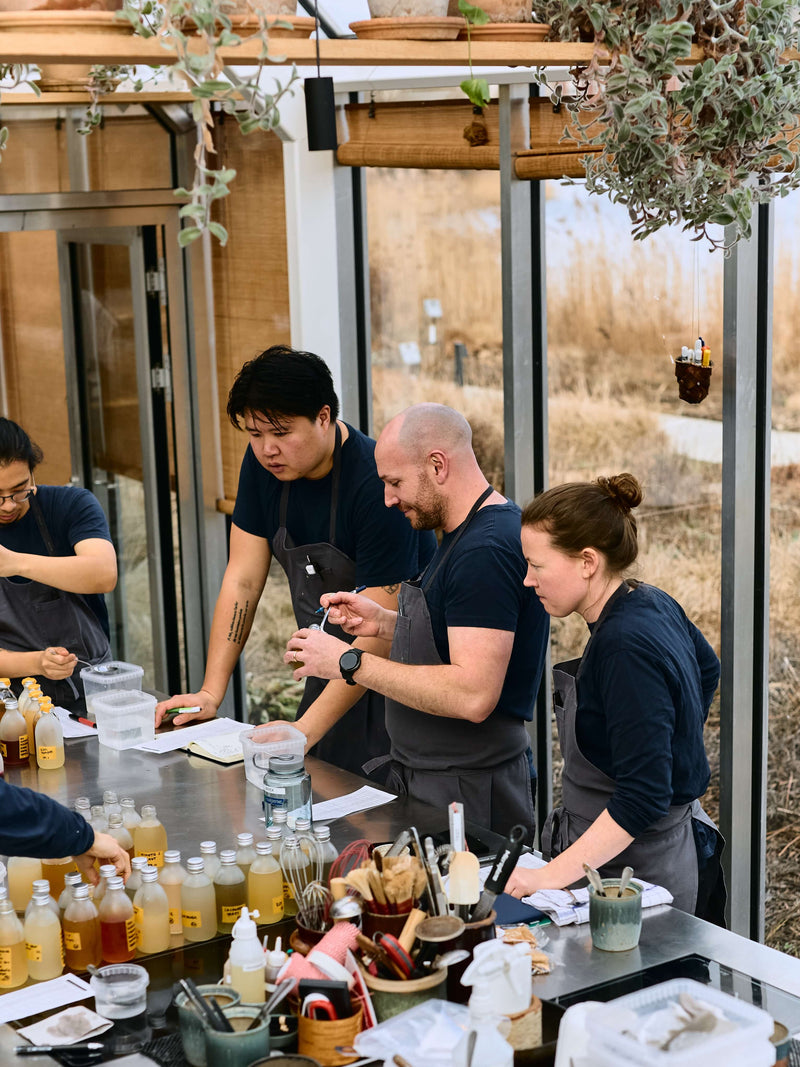 Group of people in a kitchen setting with various bottles and ingredients on a table.